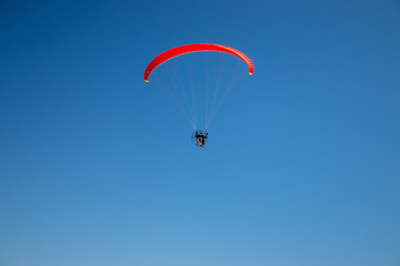 Paramotor (powered paraglider) with red parachute flying in blue sky. Back view.