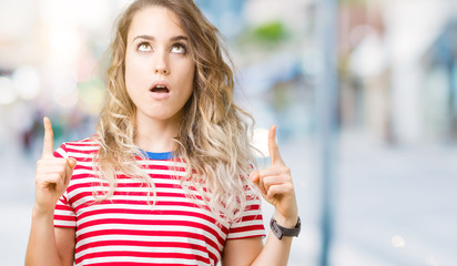 Beautiful young blonde woman over isolated background amazed and surprised looking up and pointing with fingers and raised arms.