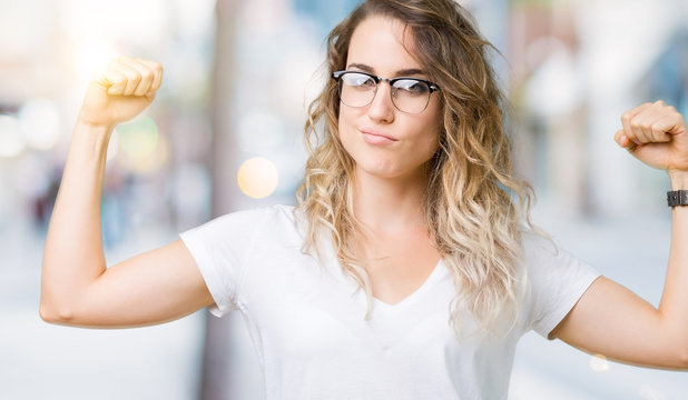 Beautiful young blonde woman wearing glasses over isolated background showing arms muscles smiling proud. Fitness concept.