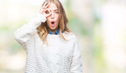 Beautiful young blonde woman wearing winter sweater over isolated background doing ok gesture shocked with surprised face, eye looking through fingers. Unbelieving expression.