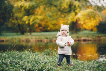 Cute little girl in a autumn park. Child in a white jacket and white hat. Kid near river