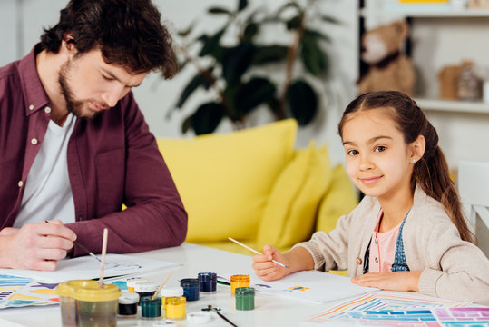 cheerful kid looking at camera while sitting near father drawing at home