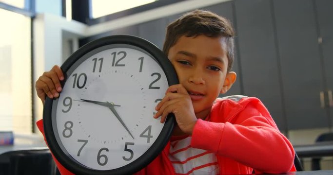 Front View Of Asian Schoolboy Holding Wall Clock At Desk In Classroom At School 4k