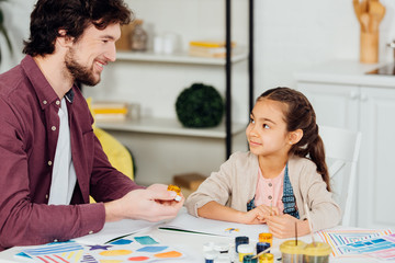 cheerful father holding gouache jar near happy daughter at home