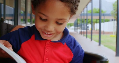 Front view of disabled African American schoolboy reading a book on wheelchair in school corridor 4k - Powered by Adobe