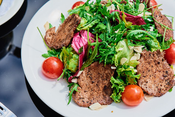 Fresh salad with vegetables and warm meat in a white plate on a dark glass table on the summer terrace. Close-up. Space