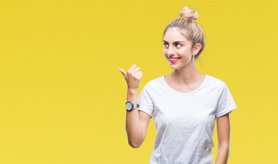 Young beautiful blonde woman wearing white t-shirt over isolated background smiling with happy face looking and pointing to the side with thumb up.
