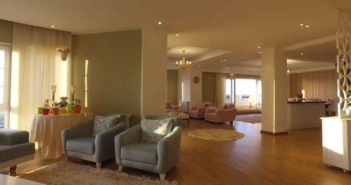 Panning shot of apartment interior   natural light coming through windows 2 with colorful flowerpot and chandelier and Persian carpet,  candlestick and  beige sofa and ,orchid dining table and kitchen