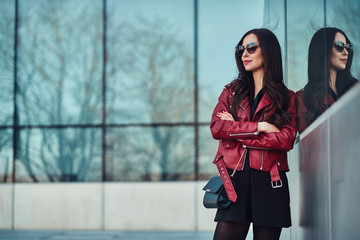 Attractive smiling woman in red jacket and sunglasses is posing near glass building.