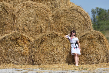 Naklejka na ściany i meble A young brunette girl in a white dress and glasses, against a background of a large haystack from round bales.