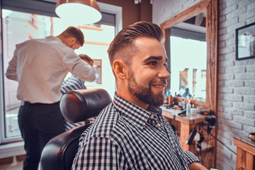 Attractive groomed man is sitting while waiting for a barber at busy barbershop. He is wearing checkered shirt.