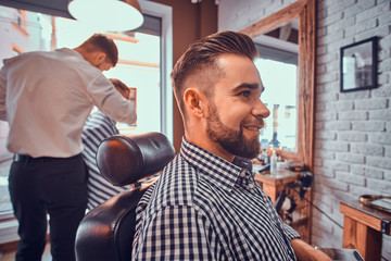 Attractive groomed man is sitting while waiting for a barber at busy barbershop. He is wearing checkered shirt.