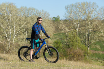 Obraz premium Cyclist in shorts and jersey on a modern carbon hardtail bike with an air suspension fork rides off-road on green hills near the forest 