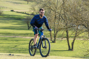 Obraz premium Cyclist in shorts and jersey on a modern carbon hardtail bike with an air suspension fork rides off-road on green hills near the forest 