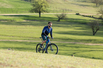 Cyclist in shorts and jersey on a modern carbon hardtail bike with an air suspension fork rides off-road on green hills near the forest	