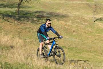 Obraz premium Cyclist in shorts and jersey on a modern carbon hardtail bike with an air suspension fork rides off-road on green hills near the forest 