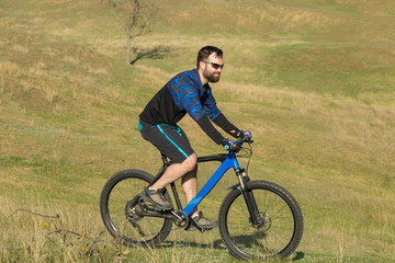 Cyclist in shorts and jersey on a modern carbon hardtail bike with an air suspension fork rides off-road on green hills near the forest	