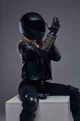 Naklejka na ściany i meble Portrait of a biker girl wearing leather racer costume and protective helmet putting on gloves in a studio on a gray background.