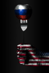 A hand with a drawn USA flag holds a fork, on which is a ball with a drawn Russia flag, a sign of influence, pressure, grip and anecxia.