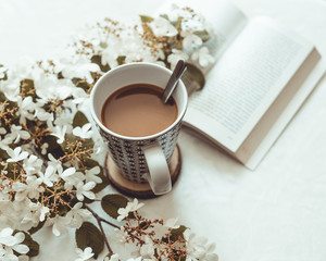 Cup of coffee with milk, with some white flowers in the blur, and an opened book