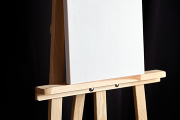 White blank cotton canvas stands on a wooden artistic easel on black curtain background. Vertical rectangular mockup canvas wrapped on stretcher bar