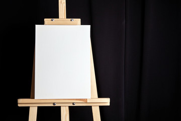 White blank cotton canvas stands on a wooden artistic easel on black curtain background. Vertical...