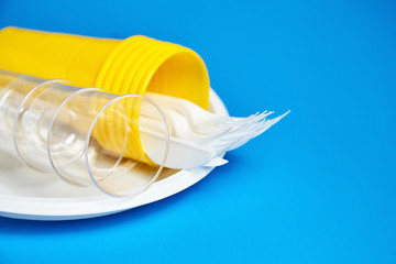 Disposable plastic tableware on blue background. Cutlery, white plate, forks, knives, yellow and transparent plastic glasses