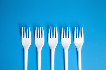 A row of lying white forks on blue background, top view. Disposable plastic tableware