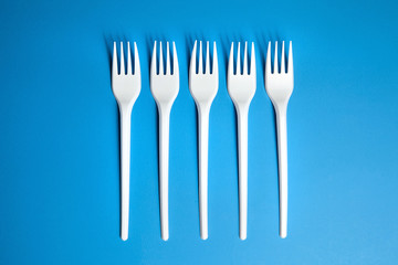 A row of lying white forks on blue background, top view. Disposable plastic tableware