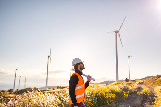 Giovane Ingegnere Aerospaziale con caschetto bianco e gilet arancione, sta controllando il progetto sul foglio di una turbina di un impianto eolico in montagna. Concetto ingegneria ed energia pulita