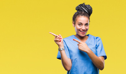 Young braided hair african american girl professional nurse over isolated background smiling and looking at the camera pointing with two hands and fingers to the side. - obrazy, fototapety, plakaty