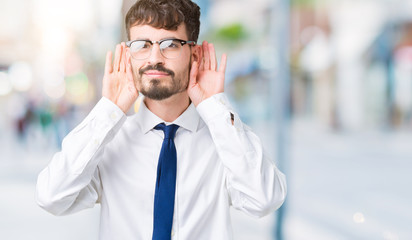 Young handsome business man wearing glasses over isolated background Trying to hear both hands on ear gesture, curious for gossip. Hearing problem, deaf