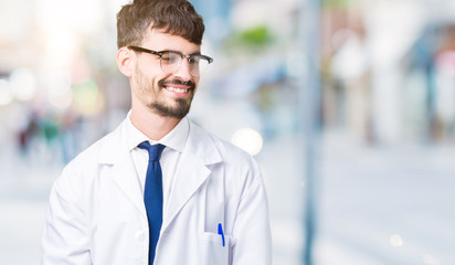 Young professional scientist man wearing white coat over isolated background looking away to side with smile on face, natural expression. Laughing confident.