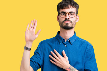 Young handsome man wearing glasses over isolated background Swearing with hand on chest and open...