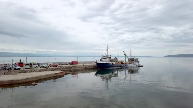 Wide, slow panning shot across Croatia harbour & medium sized fishing boat moored securely to pier. Background view of seascape & mountains under dramatic cloudy sky.