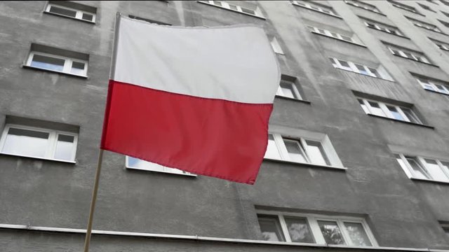 Slow motion footage of Polish flag swaying in front of grey building in Warsaw. Filmed from below.