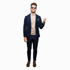 Young handsome elegant man wearing sunglasses smiling with happy face looking and pointing to the side with thumb up.