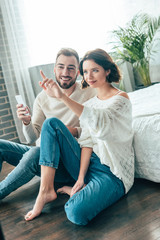 attractive brunette woman pointing with finger while sitting with handsome man holding smartphone at home
