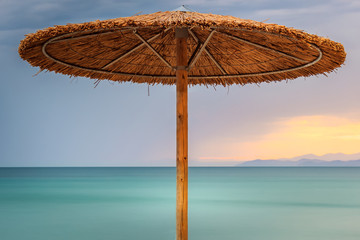 Straw beach parasol, sunset sky and silky, turquoise sea water on Paradisos beach in Neos Marmaras, Greece