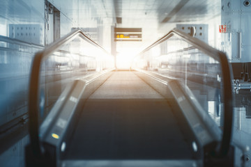 A long modern travelator indoors of an airport terminal with bright exit in front; glass corridor with a contemporary moving walkway in interior of departure or arrival area railway station depot