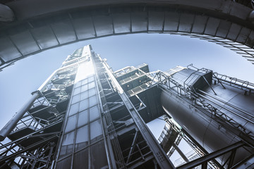 Wide-angle bottom view of a contemporary construction of an oil refinery or a modern fuel factory facility in an industrial zone, with a round bridge, plenty of pipes, iron beams, tanks, and stairs