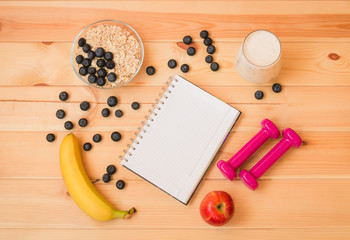 Glass of yogurt,  banana, blueberries, apple, oat flake, notebook and dumbbells on wooden background.