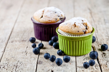 Two homemade fresh muffins on ceramic green and purple bowls with blueberries on rustic wooden table.