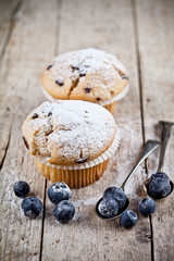 Two homemade fresh muffins with sugar powder, vintage spoons and blueberries on on rustic wooden table.