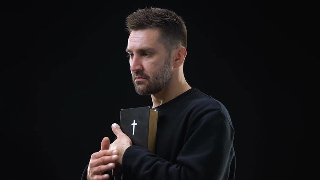 Sad parishioner holding bible closing eyes on dark background, holy scripture