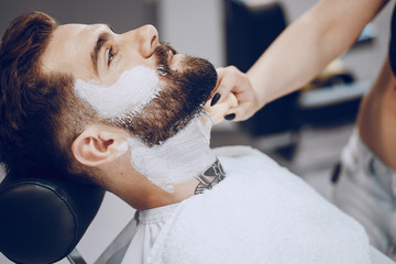 Obraz na płótnie Canvas handsome young bearded guy sitting in an armchair in a beauty salon and the girl around him smears his beard with cream