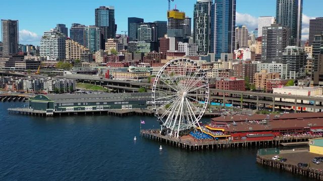 Aerial, tracking, drone shot, panning around the Seattle Great Wheel, ferris wheel and buildings and skyscrapers in the background, in downtown Seattle, on a sunny day, Washington state, USA
