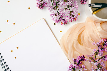 Flat lay, top view female office table desk. Workspace with paper notebook, stationery, purple lilac flowers, orange scarf, confetti on white background. Feminine desktop, minimalist style concept.