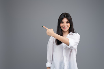 Smiling young business woman in white shirt pointing at copy space with her finger isolated over grey background