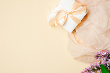 Feminine desk with silk scarf, gift box and lilac flowers on beige background. Top view, tender minimal flat lay style composition with copy space